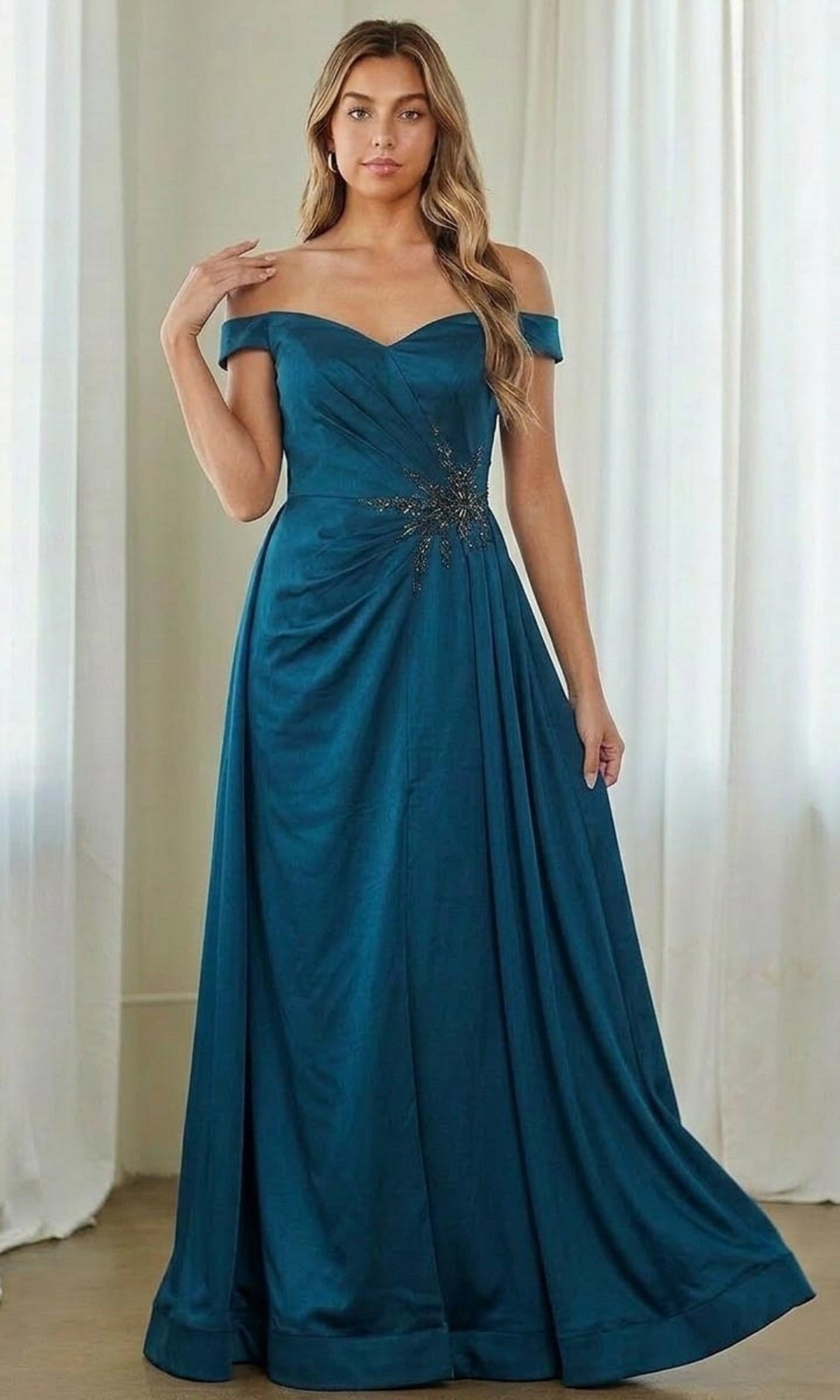 A woman stands indoors by white curtains, wearing the teal Amelia Couture 7094 long formal dress—an off-the-shoulder mother of the bride gown with a gathered waist and elegant embellishments.